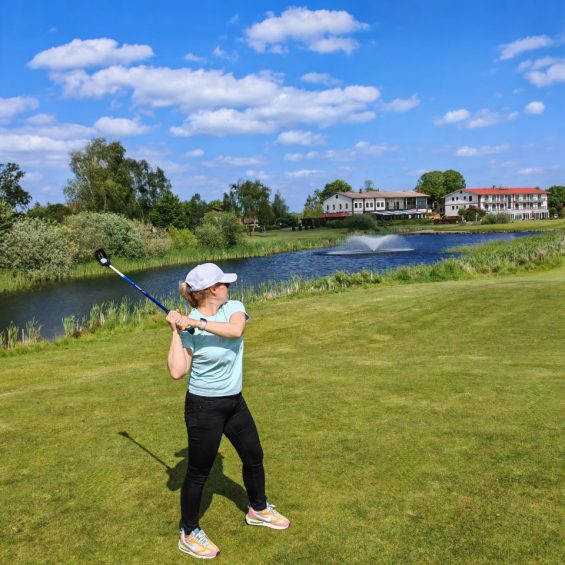 FlingGolf Spielerin auf dem Golfplatz mit FlingGolf Schlaeger im Hintergrund Wasserfontaine und Restaurant Frettwurst