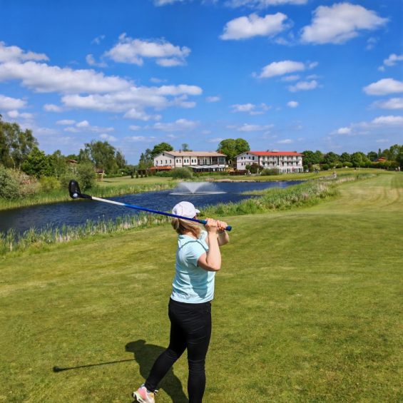 FlingGolf Spielerin auf dem Golfplatz mit FlingGolf Schlaeger. Im Hintergrund Wasserfontaine und das Restaurant Frettwurst im Golfpark Strelasund.