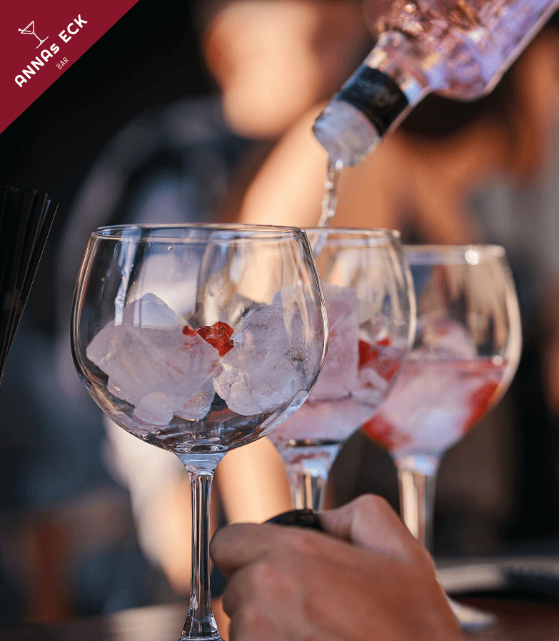 Drei gefüllte Gläser mit Eiswürfel in denen aus einer Flasche Gin eingegossen wird, eine Hand hält ein Glas fest